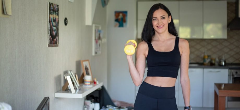 Smiling woman in activewear lifting dumbbells at home for a healthy lifestyle.